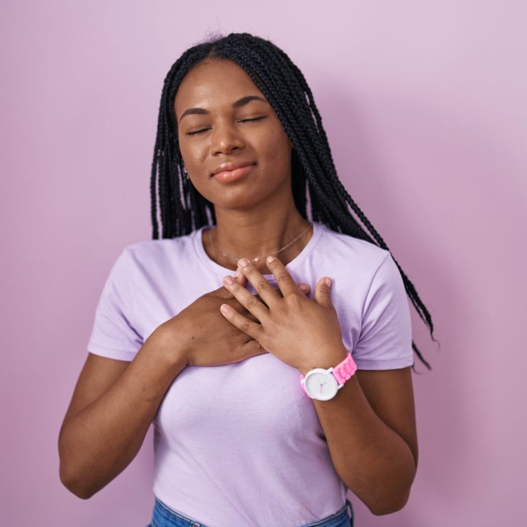 African american woman with braids standing over pink background smiling with hands on chest with closed eyes and grateful gesture on face. health concept.