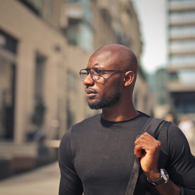 A young African male with glasses wearing a black t-shirt and a backpack in the street